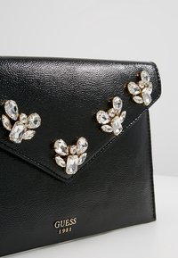 Svart texturerad läderclutch med en guldaccentuerad flap. Innehåller tre juveldekorationer och en logotyp längst ner. Rektangulär form.