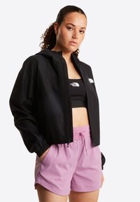 Γυναίκα με σγουρά μαλλιά φοράει μαύρο μπουφάν με φερμουάρ, μαύρο αθλητικό μπουστάκι και ροζ σορτς με κορδόνι, στέκεται με το ένα χέρι στην τσέπη.