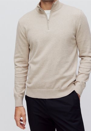 Muž v béžovém pulovru se zipem do poloviny a bílém tričku, černé kalhoty, jedna ruka v kapse na jednotném pozadí.