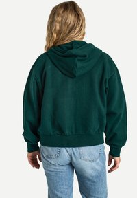 Haut en molleton court vert foncé avec capuche, poignets et ourlet côtelés. Tissu doux au toucher lisse. Porté avec un jean bleu clair.
