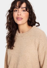 Beež neeliku särk ümmarguse kaelusega, tekstuurse kudumisega ja peente sädelevate aktsentidega. Omab pikki, lõdvaid varrukaid ja vabalt istuvat lõiget.