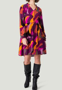 Robe violette avec un motif abstrait orange, rose et marron, dotée de manches longues et d'une taille froncée, associée à des bottes noires montantes.