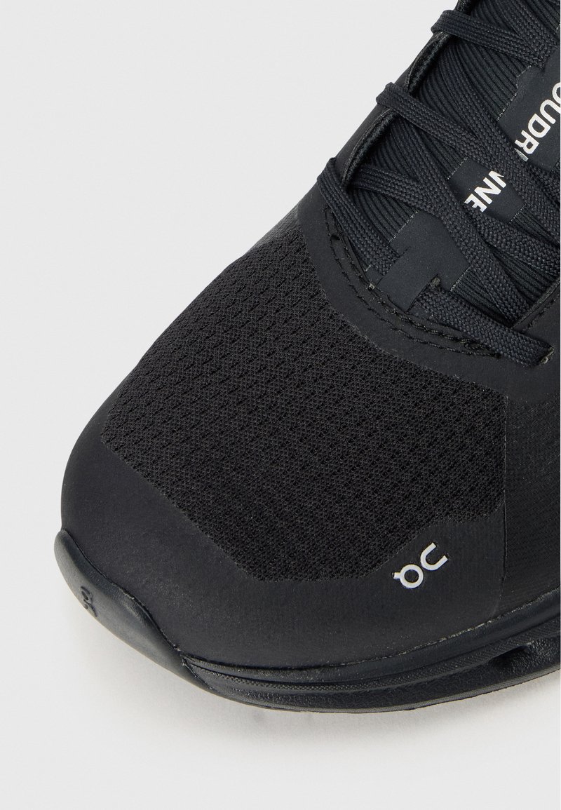 Zapatilla deportiva negra con parte superior de malla texturizada, superposiciones sintéticas lisas y suela de goma. Logotipo notable en el costado.