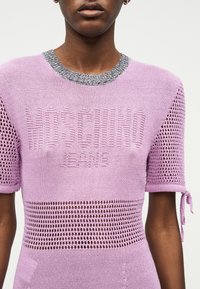 Κοντινό πλάνο ατόμου που φοράει λιλά πλεκτό μπλουζάκι κοντομάνικο με τμήματα από δίχτυ και την επιγραφή "MOSCHINO JEANS" ανάγλυφη στο μπροστινό μέρος.