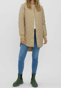 Beige, gestepptes Langarm-Mantel mit Knopfverschluss, gerippten Bündchen und zwei seitlichen Taschen. Getragen über blauen Jeans und grünen Stiefeletten.