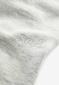 Helle graue Baumwollstoff mit gesticktem Text "self" in einer leicht dunkleren Nuance, der eine glatte, weiche Textur zeigt.
