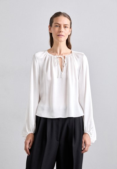 Blouse blanche à manches longues avec un col froncé et une fermeture à nouer, présentant une texture lisse et une coupe décontractée. Associée à un pantalon noir.