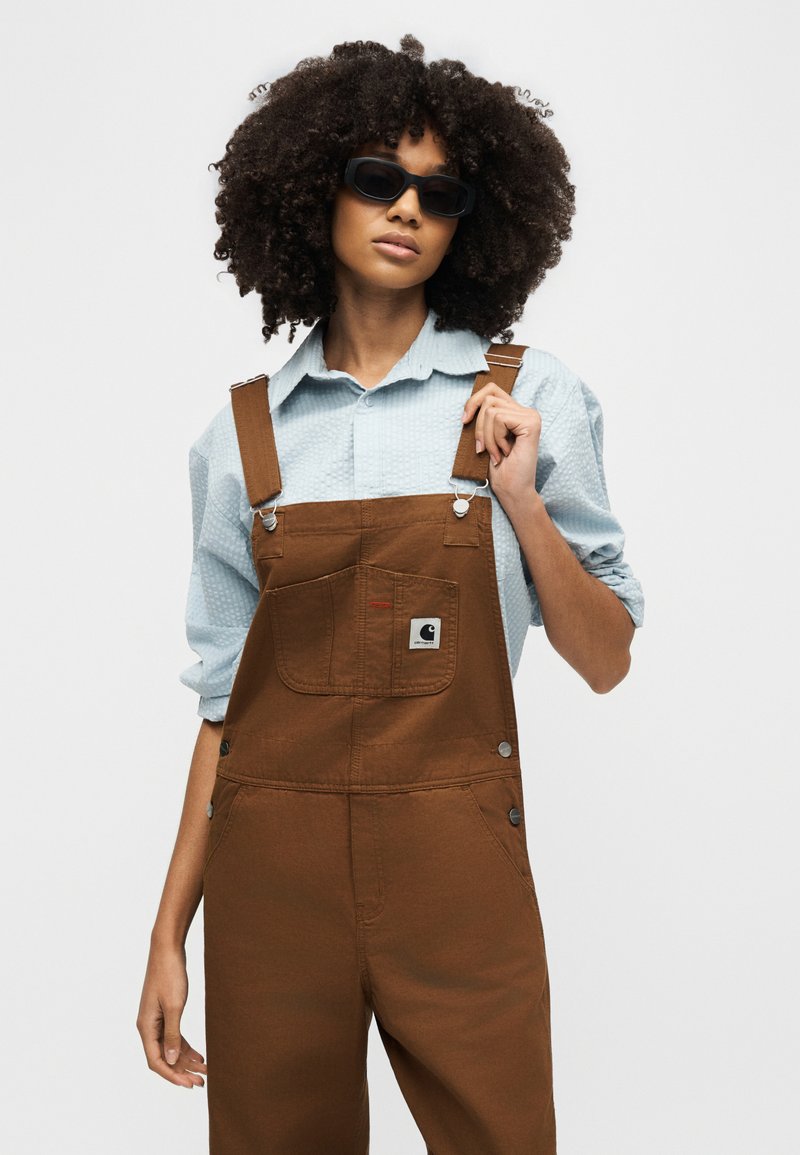 Woman with curly hair wearing black sunglasses, light blue collared shirt, and brown overalls against a plain background.