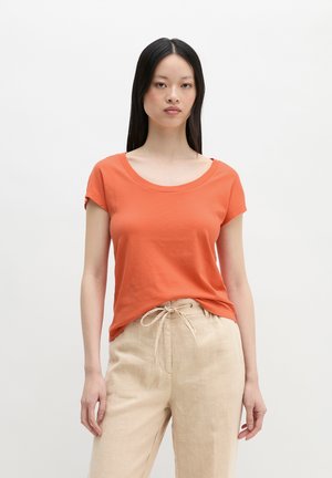 Jonge vrouw met lang zwart haar, die een oranje T-shirt met korte mouwen en beige broek met trekkoord draagt, staand tegen een effen witte achtergrond.