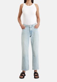 Mujer con una blusa blanca sin mangas, jeans rectos de cintura alta en azul claro, sandalias negras con hebilla, de pie con las manos en los bolsillos.