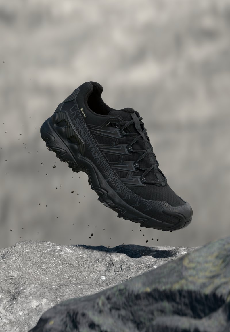 Zapato deportivo negro con parte superior texturizada, sistema de cordones y lados con patrones; cuenta con una suela gruesa y resistente y una apariencia flotante sobre una roca.