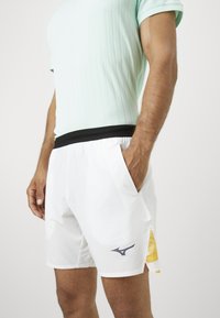 Mann in weißen Sportshorts mit schwarzem Bund und gelben Netz-Seiteneinsätzen, der mit einer Hand in der Tasche steht.