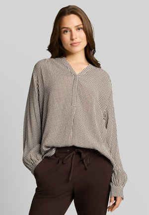 Jonge vrouw met los bruin haar, die een zwart-wit patroon blouse met lange mouwen en donkerbruine broek met koordsluiting in de taille draagt.