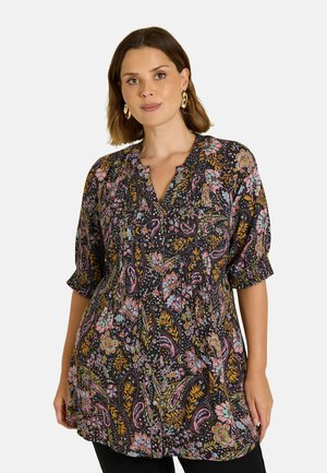 Mujer con una túnica negra con un patrón floral de cachemira en rosa, azul y mostaza y mangas abullonadas hasta los tres cuartos, de pie sobre un fondo blanco.
