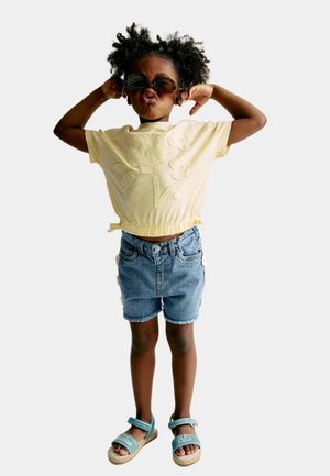 Jeune enfant aux cheveux bouclés portant des lunettes de soleil, une chemise jaune à fleurs, un short en jean et des sandales bleues, posant avec les mains derrière la tête.