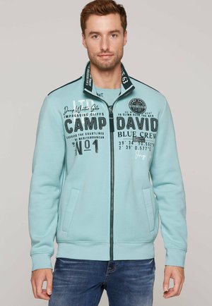 Sudadera con cremallera de color azul claro, que presenta gráficos de texto en negro, dos bolsillos laterales y puños acanalados. Confeccionada con una tela suave y texturizada.