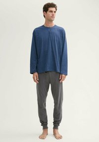 Langetermijn mouwen blauwe shirt met een ronde hals en grijze joggerbroeken, beide gemaakt van zachte stof, gedragen door een staande mannelijke figuur.