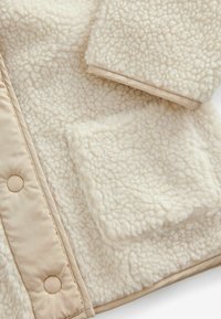 Nepbont jasje in roomkleur met een textuur en een fluffige afwerking. Bevat een beige accent en een voorzijde zak met klep. Inclusief gladde drukknoopjes.