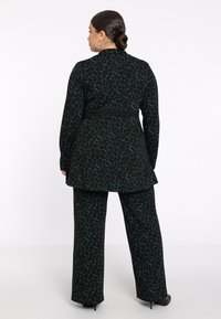 Vrouw met donker haar in een lage bun, gekleed in een lange zwarte en groene gepatterniseerde jas en bijpassende wijde broek, staande tegen een witte achtergrond.