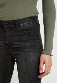 Jeans vaqueros negros ajustados, con cierre de botón y cremallera, bolsillos delanteros y textura ligeramente desgastada. Combinados con una camiseta verde acanalada.
