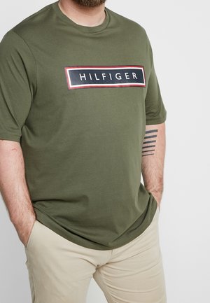 Olive green t-shirt with short sleeves, round neckline, and a large rectangular logo featuring “HILFIGER” in bold black and white.