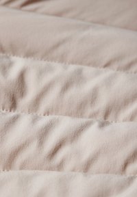 Gepolsterter Stoff in sanftem Beige mit sichtbarer Struktur und Nähten; zeigt subtile, plüschige Falten und eine glatte Oberfläche, die Wärme und Komfort vermittelt.