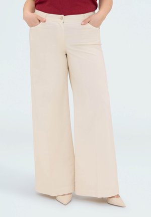 Personne portant un pantalon beige taille haute à jambes larges, les mains dans les poches et des chaussures beige pointues, associé à un haut rouge.