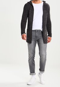 Długi, ciemnoszary sweter z dzianiny z kapturem, otwarty z przodu, z dwiema przednimi kieszeniami, w zestawie z białą koszulą i szarymi dżinsami. Białe sneakersy.