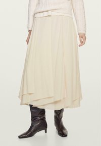 Cremfarbener, knielanger Rock aus leichtem Stoff mit geschichteter Gestaltung. Kombiniert mit glänzenden schwarzen Stiefeletten. Sanfte Textur und weicher Fall.