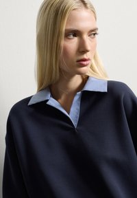 Mujer rubia con una blusa azul marino y un cuello de cuadros azul claro, mirando hacia la derecha frente a una pared blanca lisa.