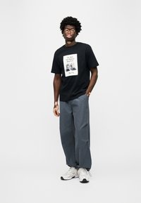 Μαύρο μπλουζάκι με γραφικό σχέδιο στο μπροστινό μέρος, συνδυασμένο με χαλαρά γκρι παντελόνια και λευκά αθλητικά παπούτσια. Το μοντέλο στέκεται με τα χέρια στις τσέπες.