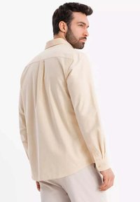 Beige Langarmhemd mit einem Rückenschlitzdetail, Knopfmanschetten und einem spitzen Kragen, aus einem glatten Stoff gefertigt.