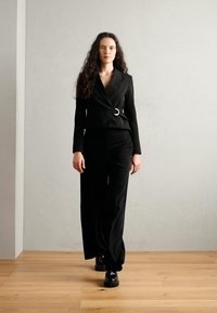 Black tailored suit with wide-leg trousers and a double-breasted blazer, featuring a circular belt buckle detail and textured fabric.