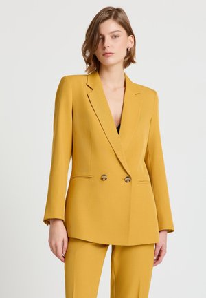 Giovane donna con blazer doppiopetto giallo senape e pantaloni abbinati, in piedi davanti a uno sfondo chiaro e uniforme.