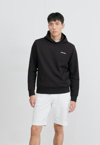 Černá mikina s kapucí a přední klokaní kapsou, úzkými manžetami a šedým logem Calvin Klein. V kombinaci s bílými šortkami s logem.