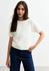 Pull court blanc et moelleux à manches courtes avec un col rond, associé à un jean bleu foncé. Texture douce, coupe décontractée et sans motifs ni accents visibles.