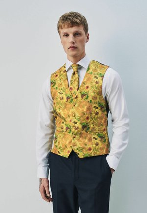 Jeune homme portant une chemise blanche, un pantalon foncé, un gilet jaune vif à motif floral et une cravate assortie sur un fond uni.