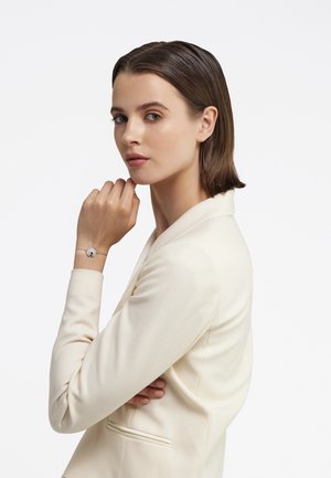 Jauna sieviete krēmveida žaketē rāda sudraba rokassprādzi ar apaļu piekariņu uz vienkārša balta fona.