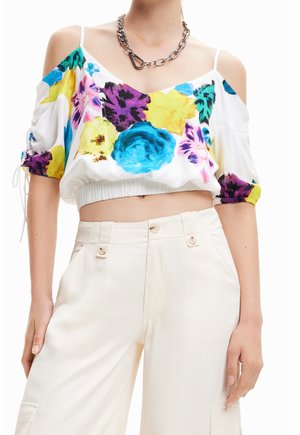 Femme portant un crop top blanc à motif floral avec des manches tombantes et un pantalon crème taille haute, accessoirisé d’un collier chaîne épais.