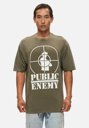 Camiseta oversized verde oliva, de material de algodón, presenta un gran gráfico en blanco con un blanco y el texto "PUBLIC ENEMY" en la parte inferior.