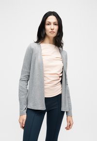 Γυναίκα με σκούρα μαλλιά φοράει ανοιχτό ροζ τοπ με ρυτιδώσεις, ανοιχτή γκρι ζακέτα και σκουρόχρωμο στενό παντελόνι, μπροστά σε απλό λευκό φόντο.