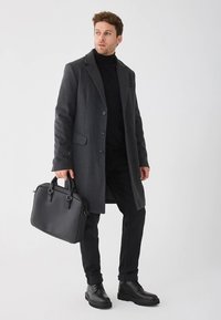 Soprabito in lana grigio scuro con rever a incavo, chiusura con bottoni e tasche laterali, abbinato a un dolcevita nero e pantaloni neri, con una borsa in pelle nera in mano.