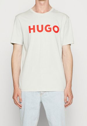 Homme portant un t-shirt gris clair à manches courtes avec le texte rouge audacieux "HUGO" et un jean denim bleu clair, debout devant un fond uni.