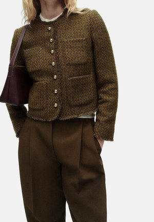 Femme portant une veste texturée marron à boutons avec quatre poches avant et un pantalon large marron assorti, portant un sac à bandoulière marron foncé.