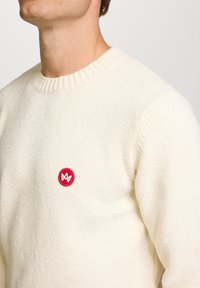 Kremfarget genser med strukturert strikk, rund halslinning og en liten rød rund merkelapp med en logo på venstre bryst.