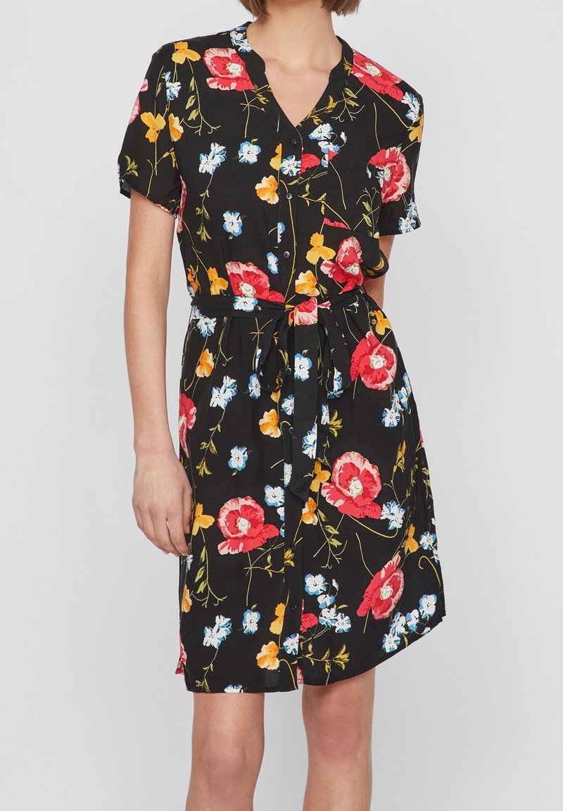Femme portant une robe noire à manches courtes à fleurs rouges, jaunes et blanches, boutonnée devant et avec une ceinture nouée à la taille.