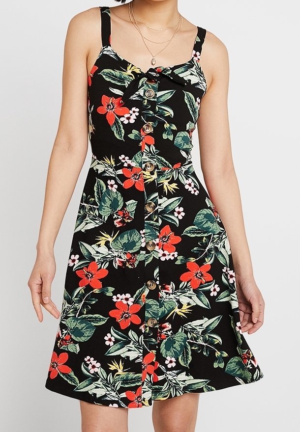 Femme portant une robe noire sans manches avec imprimé floral rouge et blanc, boutons à l'avant, et un petit détail de nœud à l'encolure.