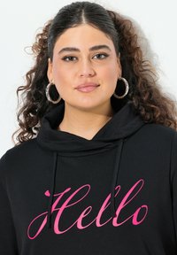 Sudadera negra con capucha que presenta un gráfico en cursiva color rosa que dice "Hola" en el frente. El tejido parece suave y liso, con un ajuste relajado.