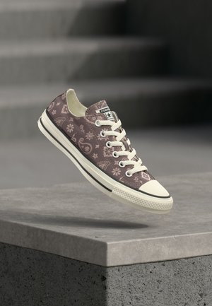 Χαμηλό sneaker Converse με μπεζ σχέδιο παϊέτας, λευκά κορδόνια και λαστιχένια σόλα που αιωρείται πάνω από ένα τσιμεντένιο χείλος.