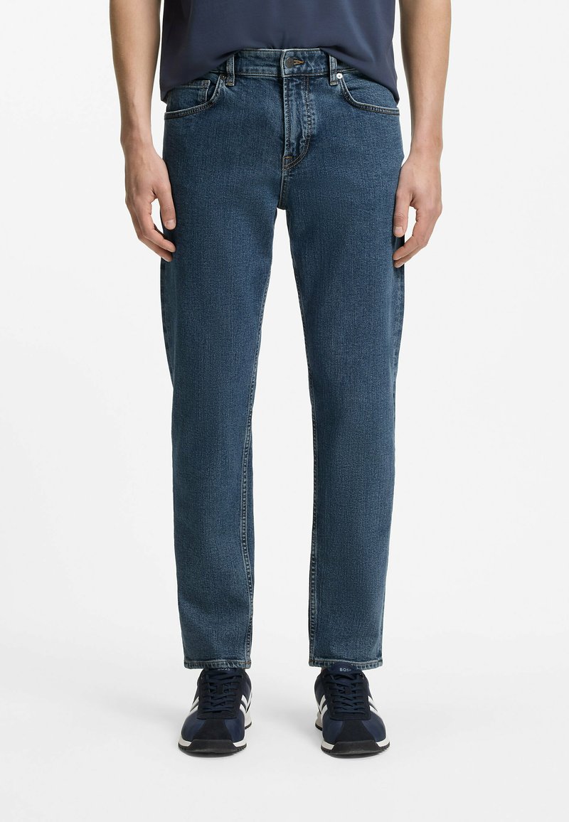 Homme portant un jean en denim bleu coupe droite, des baskets bleu marine et une chemise bleu foncé, debout devant un fond blanc uni.